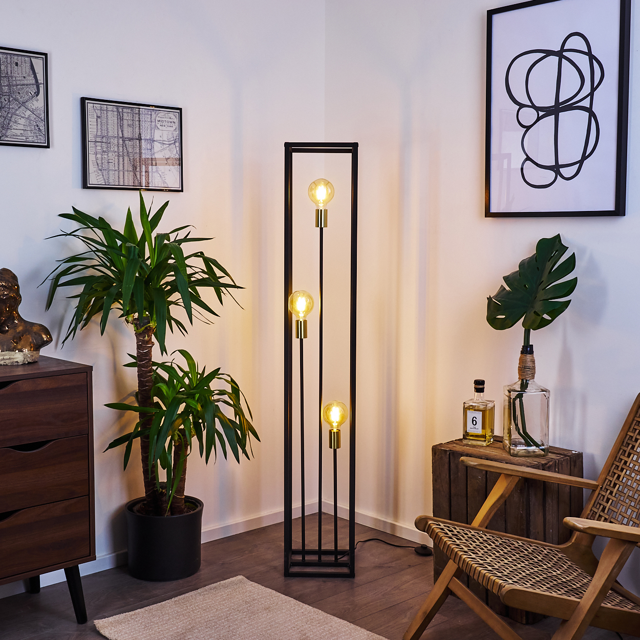 Ein Raum mit einer schwarzen Stehlampe mit drei Glühbirnen, einer Pflanze, Kunst, einer Holzkiste, einem Stuhl und einem Schrank.