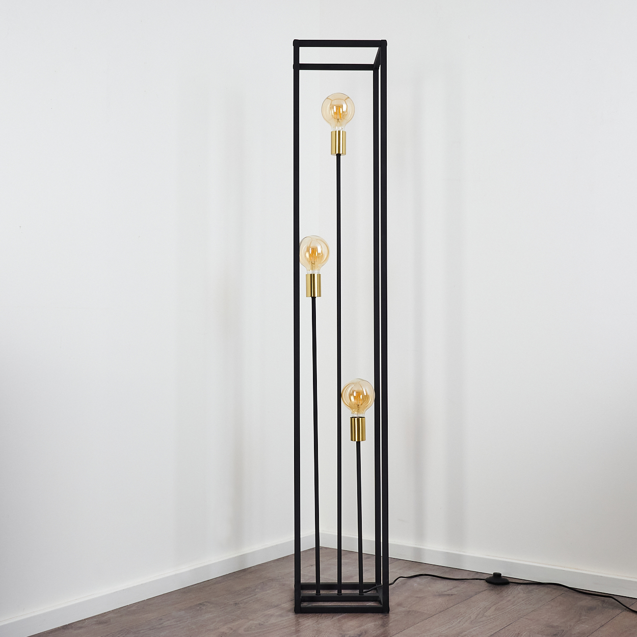 Eine schwarze Metall-Stehlampe mit drei leuchtenden Glühbirnen. Sie steht vor einer weißen Wand und einem Holzboden.