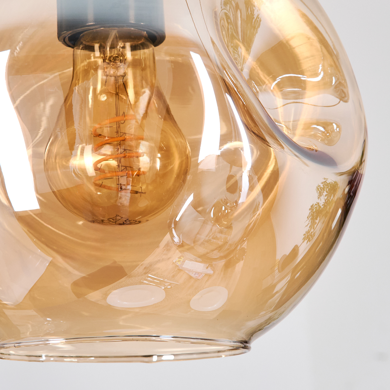 Nahaufnahme einer Glasleuchte. Eine Wendelglühbirne ist in bernsteinfarbenem, unregelmäßig geformtem Glas eingeschlossen. Weißer Hintergrund.