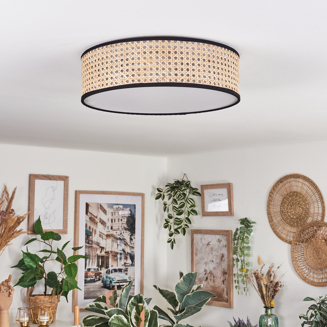 Eine Deckenlampe aus geflochtenem Rattan. Darunter zieren gerahmte Bilder und Pflanzen eine weiße Wand. Der Raum hat eine gemütliche, böhmische Atmosphäre.