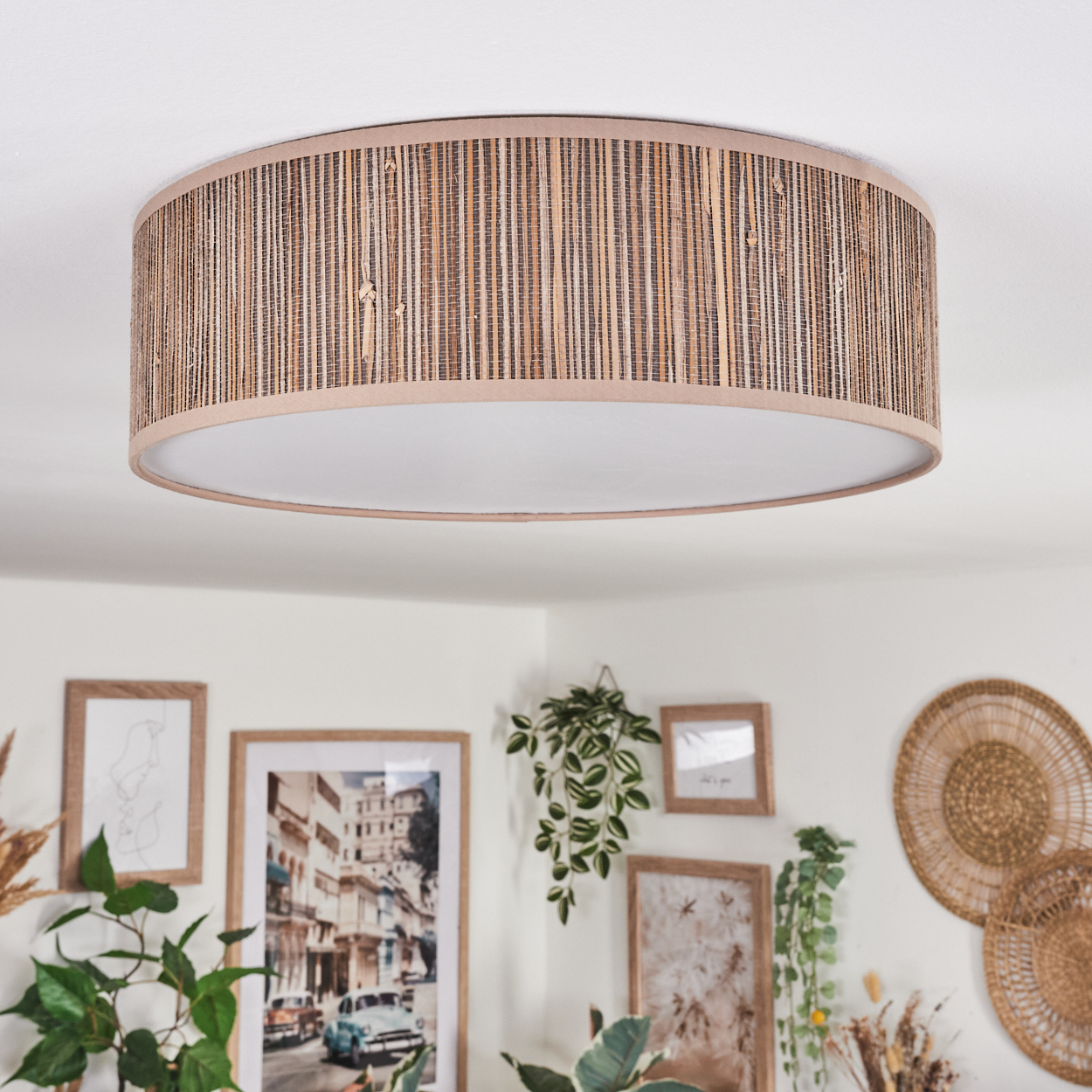 Eine Deckenlampe mit bambusähnlichem Schirm hängt über gerahmten Bildern und Pflanzen. Der Raum hat eine neutrale, natürliche Ästhetik.