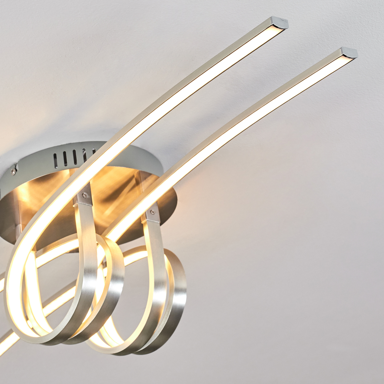Eine moderne Deckenleuchte mit gebogenen Armen aus gebürstetem Metall und integrierten LED-Leuchten. Die Leuchte ist an einer weißen Decke befestigt.
