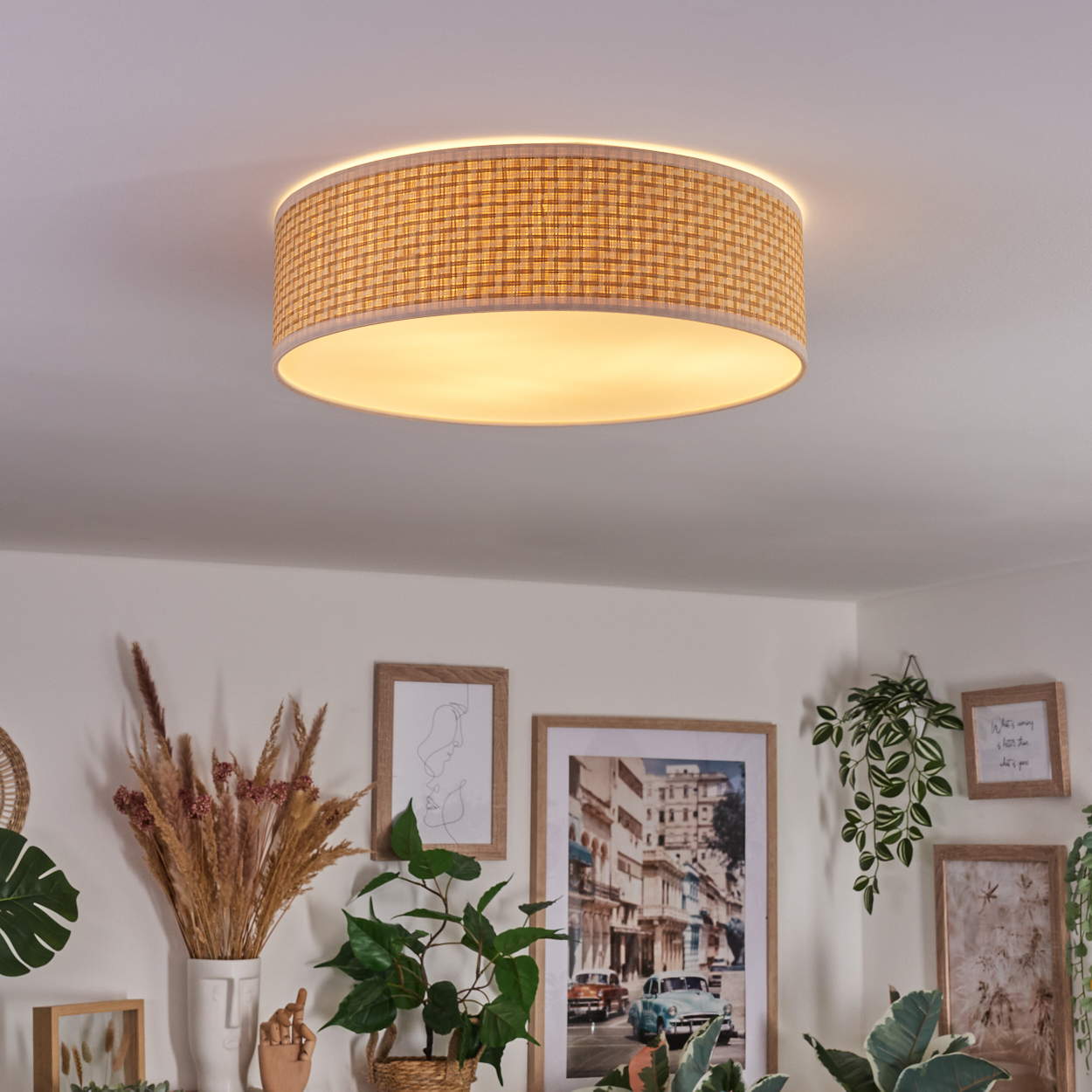 Eine Deckenleuchte mit gewebtem Lampenschirm beleuchtet einen Raum mit gerahmten Bildern und Pflanzen. Das Licht ist beige, die Wände sind weiß.