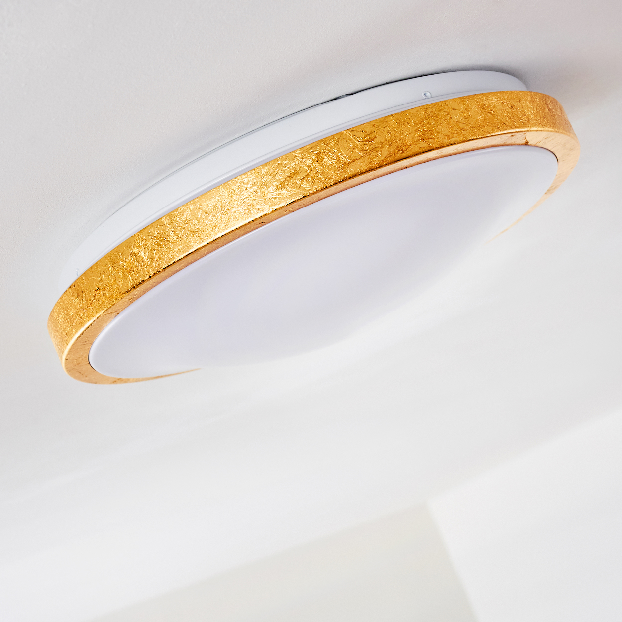 Nahaufnahme einer Deckenleuchte. Sie hat einen weißen Sockel und einen goldfarbenen, strukturierten Zierrat um das Licht.