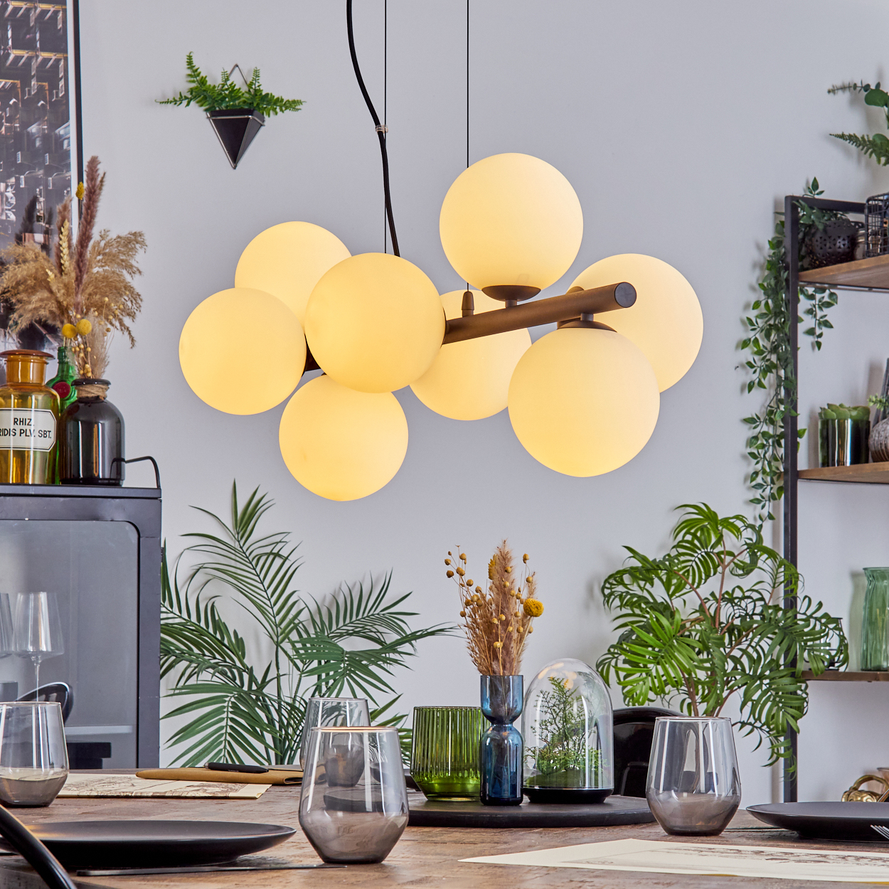 Ein Esstisch mit Kronleuchter, Pflanzen und Gläsern. Der Kronleuchter hat mehrere kugelförmige Lichter. Es gibt dekorative Pflanzen und Gläser.