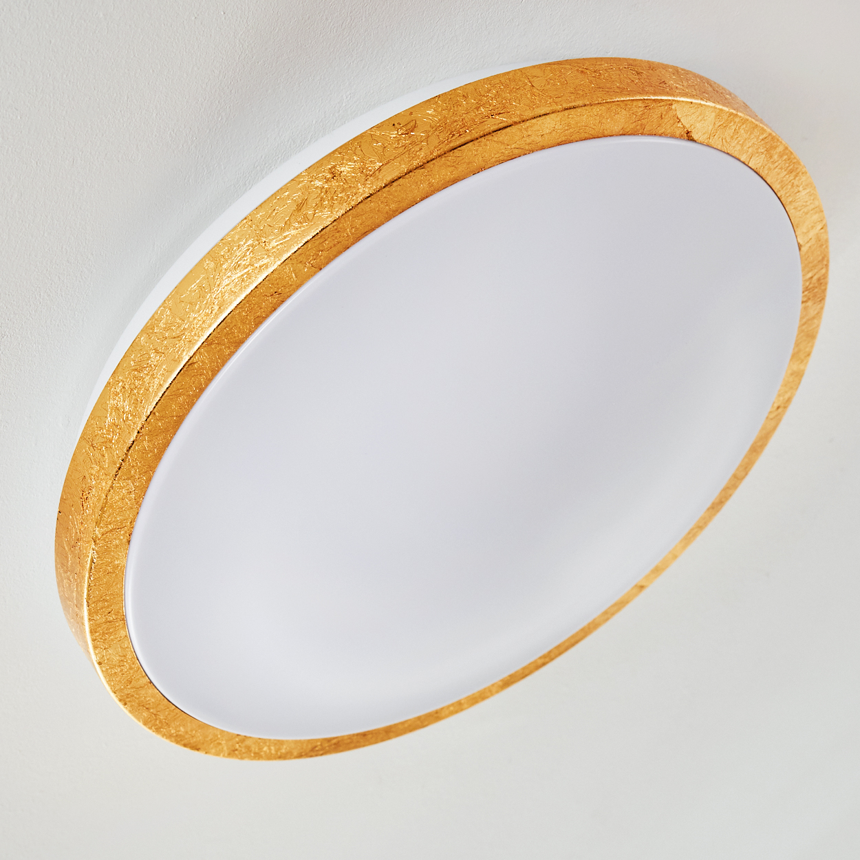 Nahaufnahme einer Deckenleuchte. Sie hat eine weiße Mitte und einen goldfarbenen, strukturierten Rahmen. Der Hintergrund ist weiß.