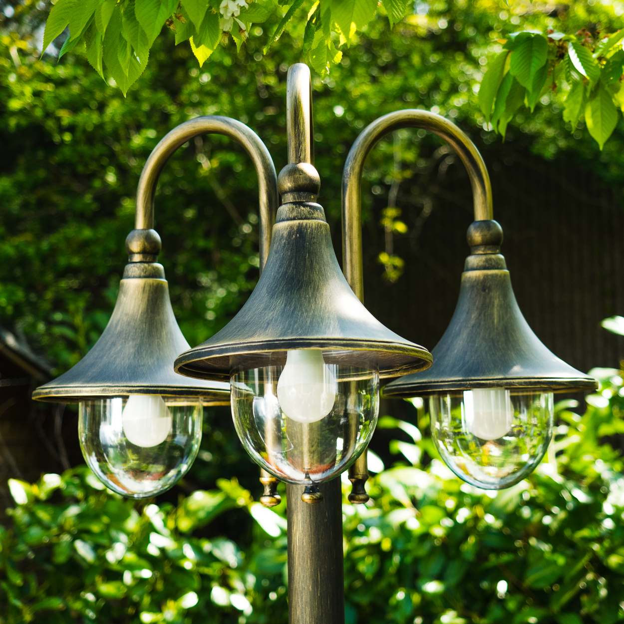 Eine verzierte Außenlampe mit drei Lichtern. Die Lampe ist bronzefarben und vor dem Hintergrund von grünem Laub und einem verschwommenen Hintergrund platziert.