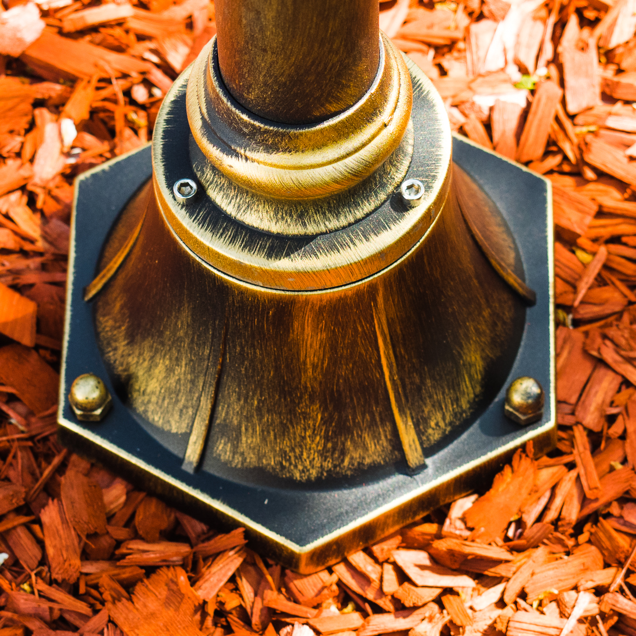 Ein bronzener Lampenfuß mit achteckiger dunkler Basis steht auf roter Mulch. Die Lampe hat ein dekoratives Design und sichtbare Schrauben.