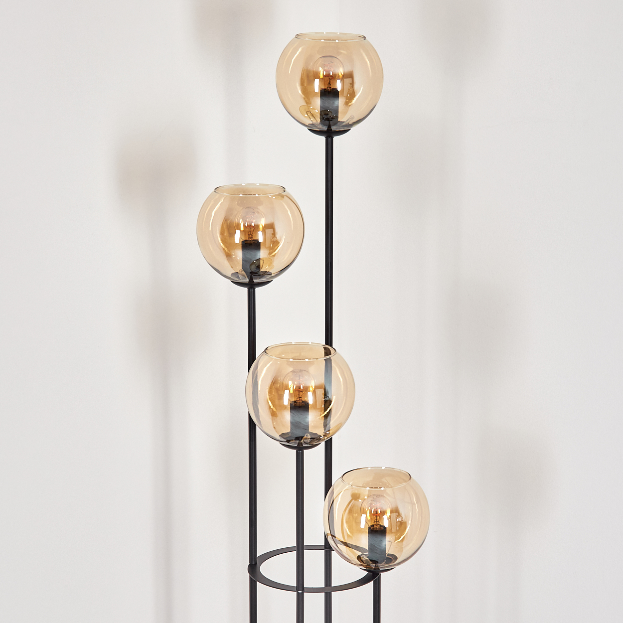 Eine moderne Stehlampe mit vier bernsteinfarbenen Glaskugeln. Das schwarze Metallgestell hat einen kreisförmigen Sockel. Der Hintergrund ist eine weiße Wand.