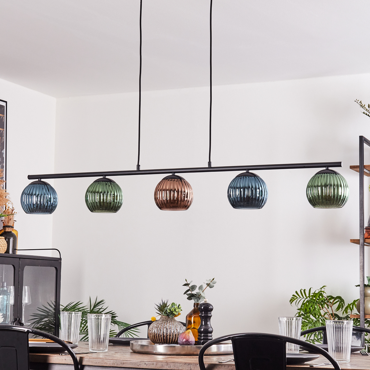 Ein Esszimmer mit Holztisch, Gläsern und einer schwarzen Leuchte mit fünf farbigen Glaskugeln.