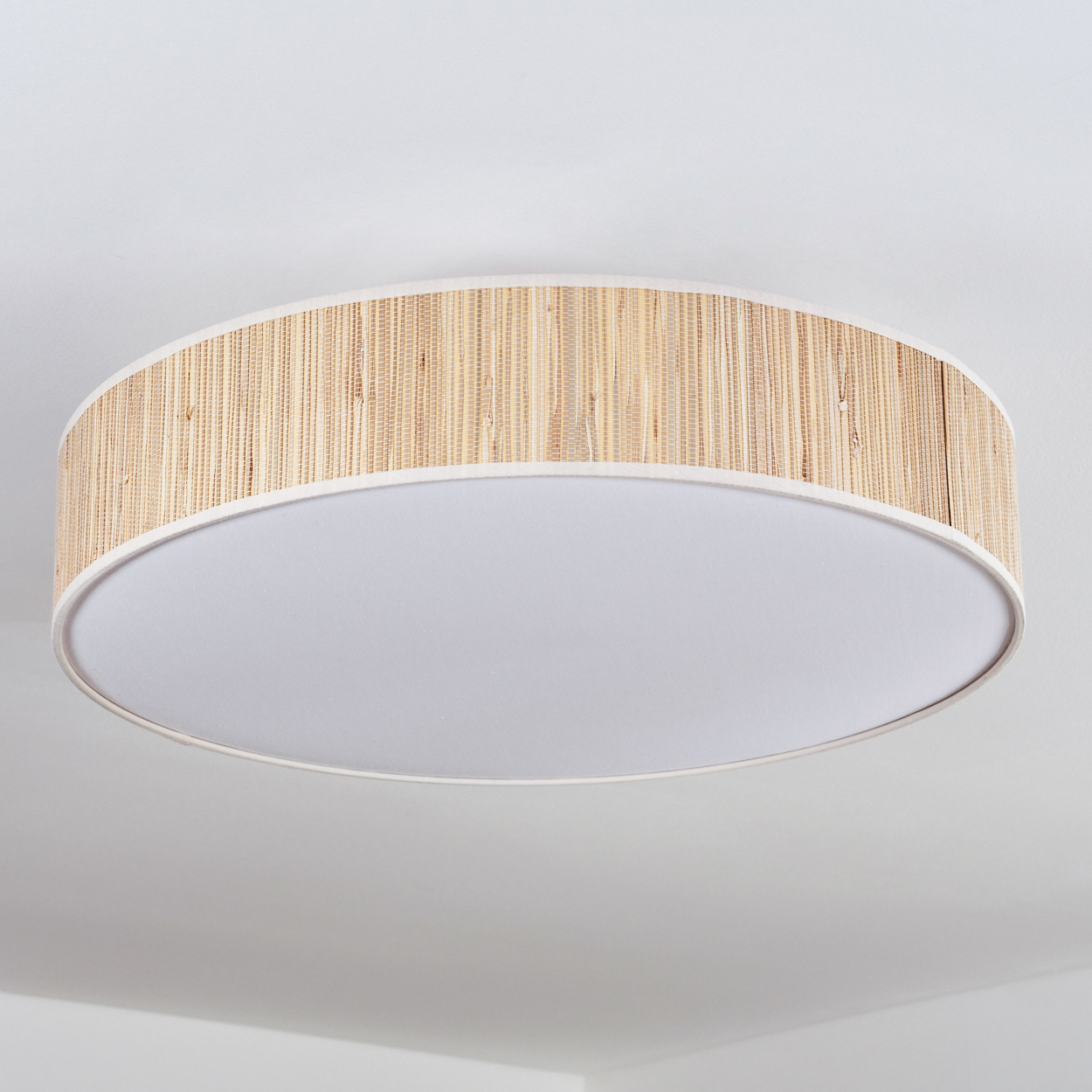 Eine runde Deckenleuchte mit einem gewebten, hellbraunen Schirm um einen weißen Diffusor. Der Hintergrund ist eine weiße Decke.