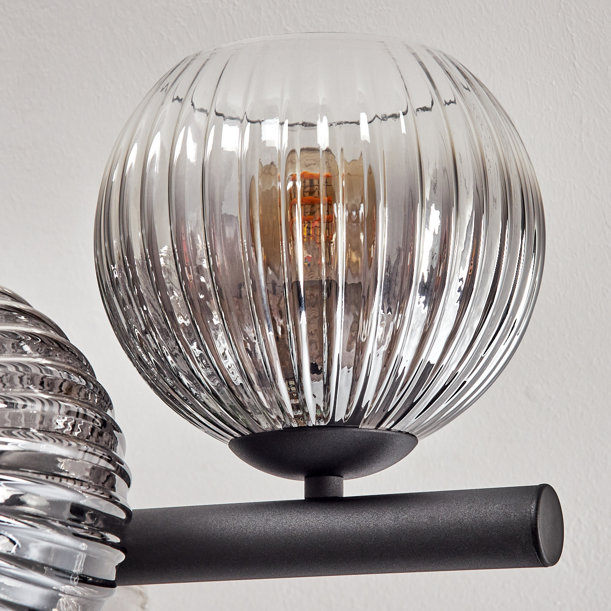Nahaufnahme einer Glaslampe mit geriffelter Oberfläche. Die Glühbirne im Inneren ist sichtbar. Die Lampe ist schwarz und an einer weißen Wand montiert.