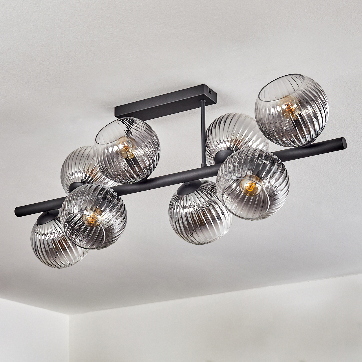 Deckenleuchte mit 8 Glaskugeln. Schwarzer Metallrahmen und Halterung. Kugeln sind spiralförmig mit Glühbirnen im Inneren. Weiße Decke.