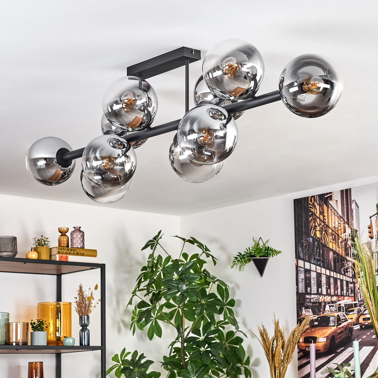 Eine moderne Deckenleuchte mit kugelförmigen Glasschirmen. Darunter ein Raum mit Dekoration, Pflanzen und einem Kunstwerk mit Stadtszene an der Wand.