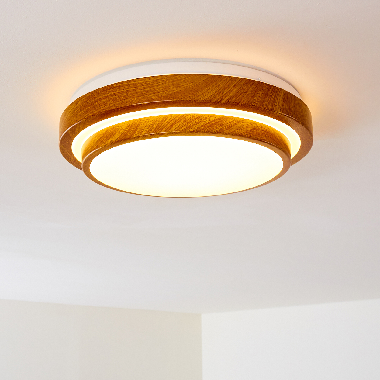 Eine moderne Deckenleuchte, holzmaseriert und weiß, mit hellem, kreisförmigem Licht in der Mitte. Sie ist an einer weißen Decke montiert.