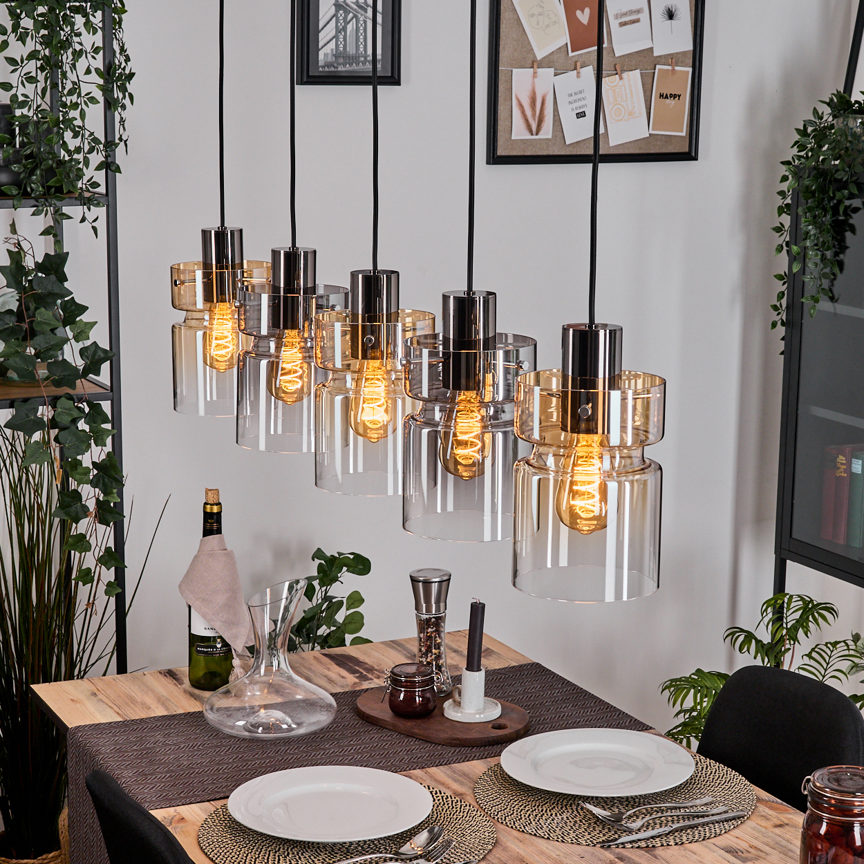 Esszimmer mit Hängelampen, einem Holztisch mit Tellern und einer Weinflasche. Pflanzen und Kunstwerke sind im Hintergrund sichtbar.
