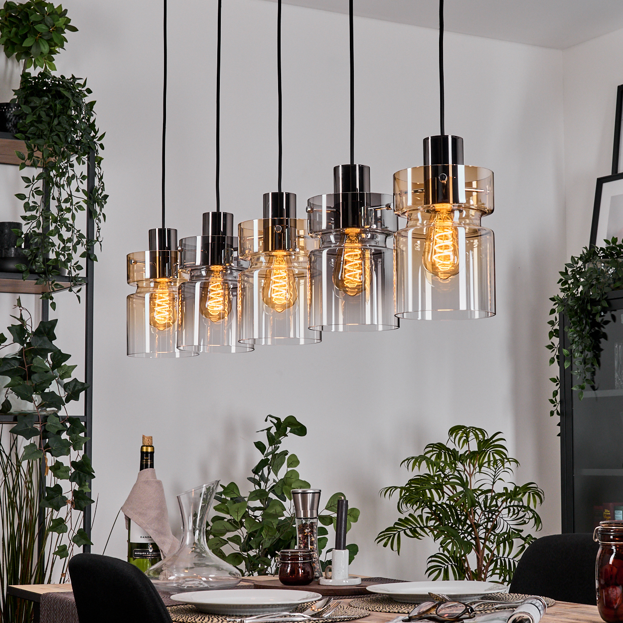 Esszimmer mit Hängelampen. Die Lampen haben Glasschirme. Ein Tisch ist mit Tellern, Gläsern und Pflanzen gedeckt.