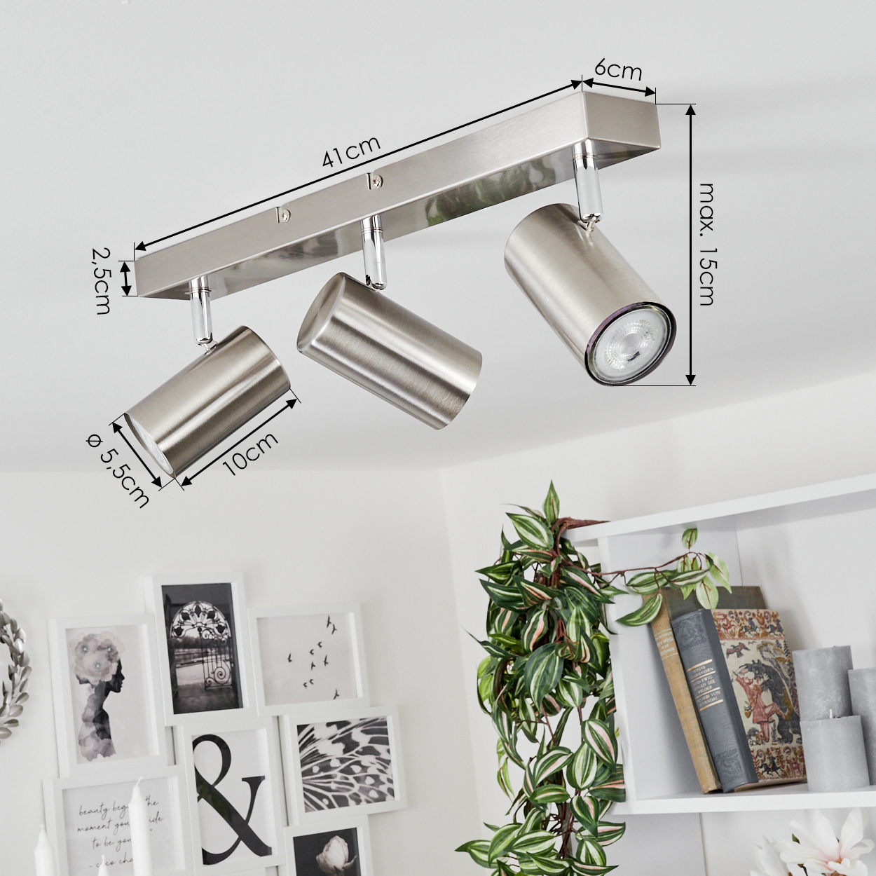 Ein moderner, silberner Deckenstrahler mit drei verstellbaren Lichtern. Abmessungen sind angegeben. Im Hintergrund sind gerahmte Kunst und ein Regal mit Pflanzen zu sehen.