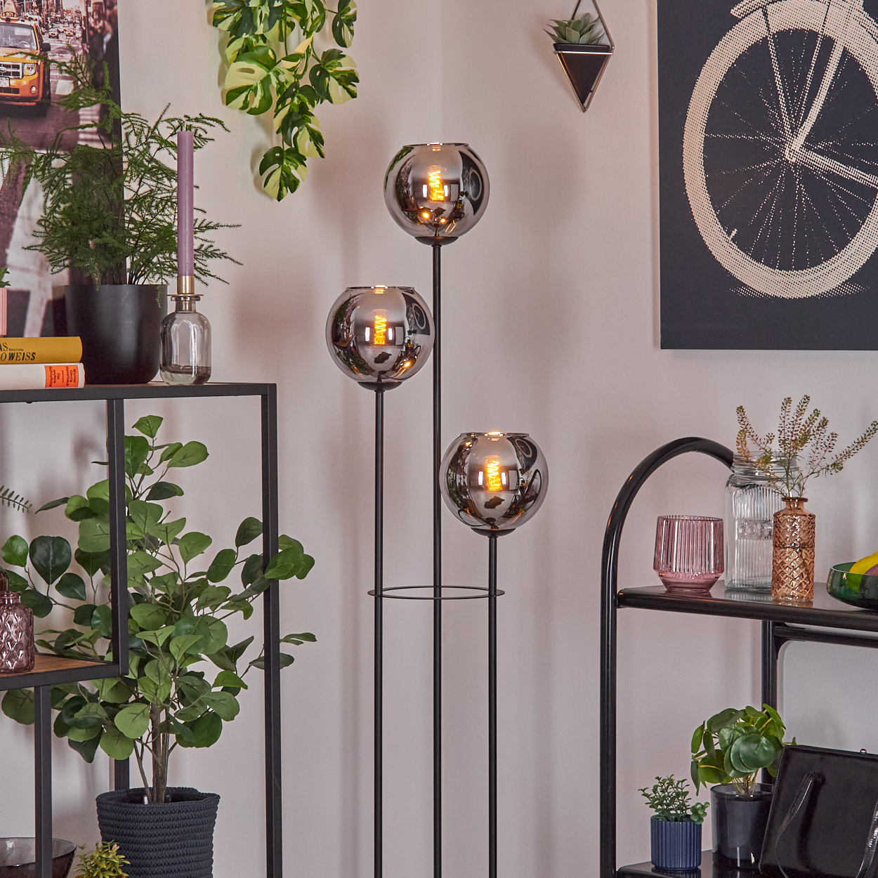 Ein Raum mit einem schwarzen Metallregal, Pflanzen und einer schwarzen Stehlampe mit drei kugelförmigen Leuchten.