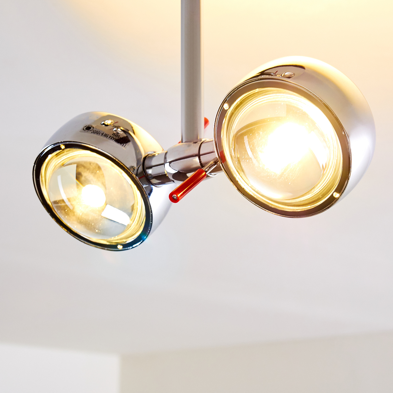 Nahaufnahme einer modernen, verchromten Deckenstrahler-Leuchte mit zwei hellen, runden Lichtern. Rote Akzente und eine weiße Decke sind sichtbar.