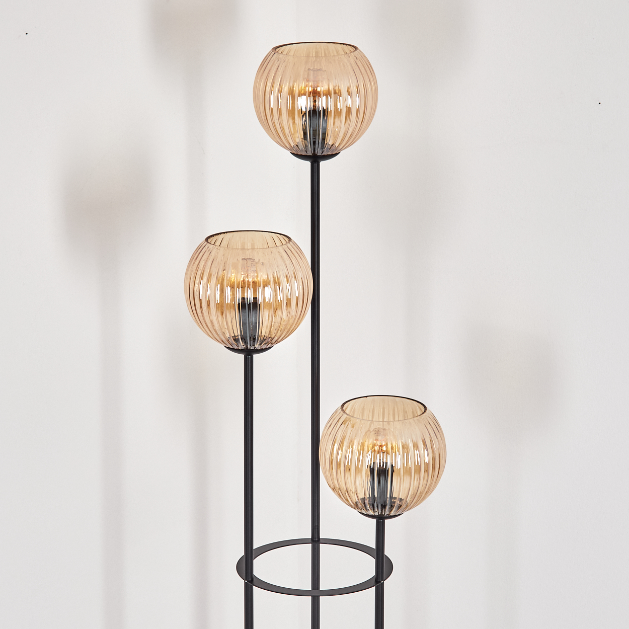 Eine moderne Stehlampe mit drei kugelförmigen, bernsteinfarbenen Glasschirmen, schwarzem Metallrahmen und weißem Hintergrund.