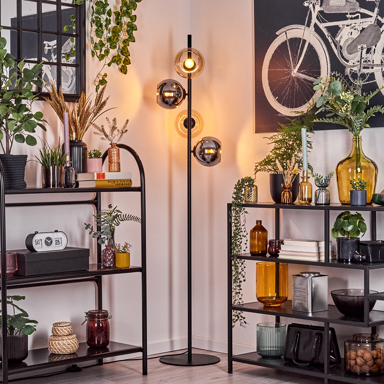 Ein Raum mit schwarzen Regalen, einer Stehlampe, Pflanzen und Dekorationsartikeln. Ein Fahrradkunstwerk befindet sich an der Wand.