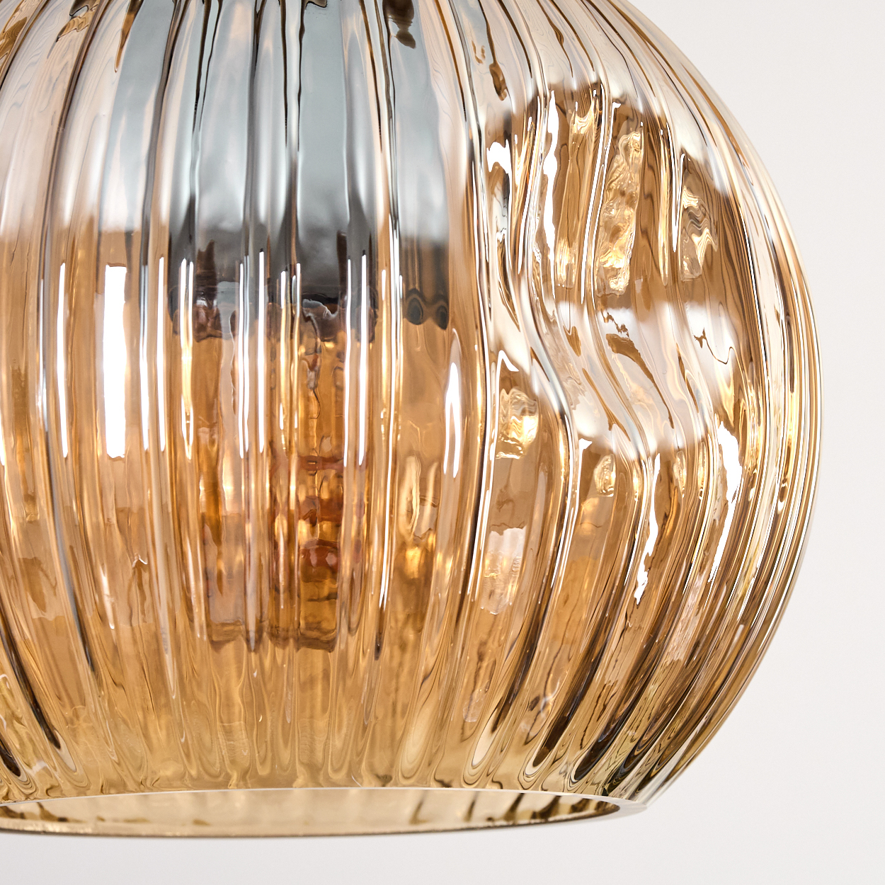 Nahaufnahme eines strukturierten, goldfarbenen Glasschirms. Vertikale Rippen erzeugen ein wellenförmiges Muster, das Licht reflektiert. Weißer Hintergrund.