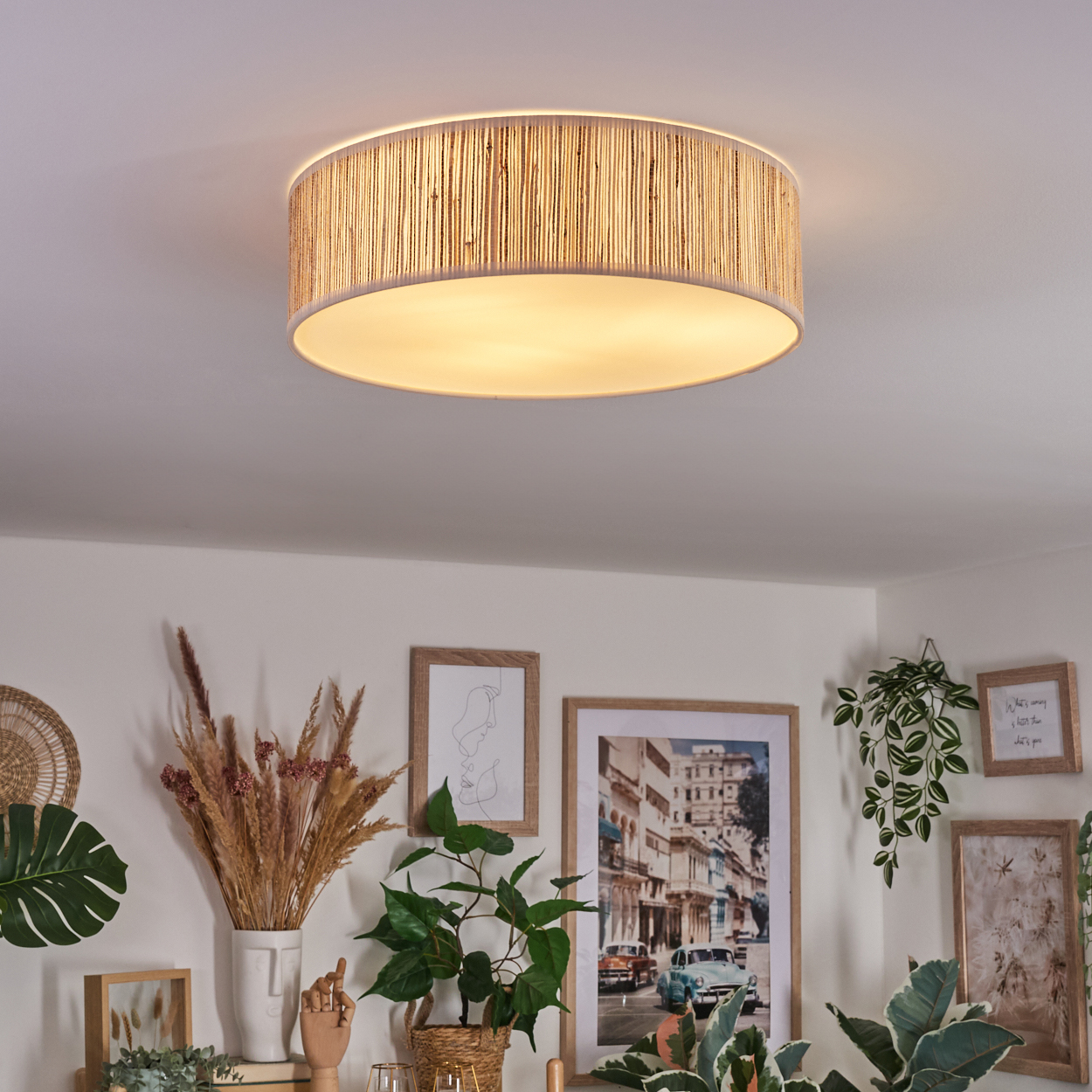Eine runde Deckenlampe mit gewebter Textur hängt über einem Raum mit gerahmten Bildern und Pflanzen. Die Lampe ist beleuchtet.