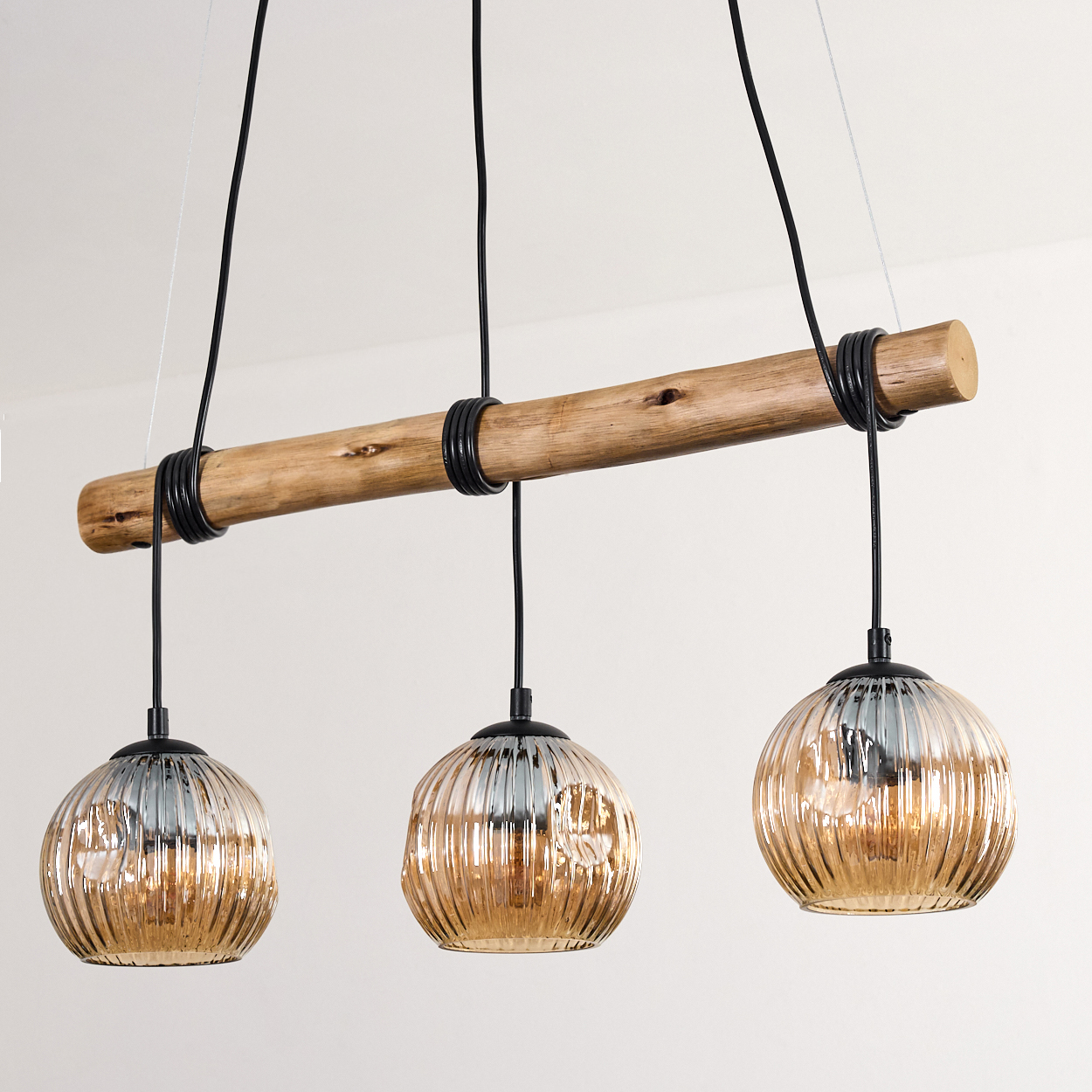 Pendelleuchte mit drei Lichtern. Die Leuchten sind rund und bernsteinfarben. Sie hängen an einem Holzbalken mit schwarzer Verkabelung vor einer weißen Wand.