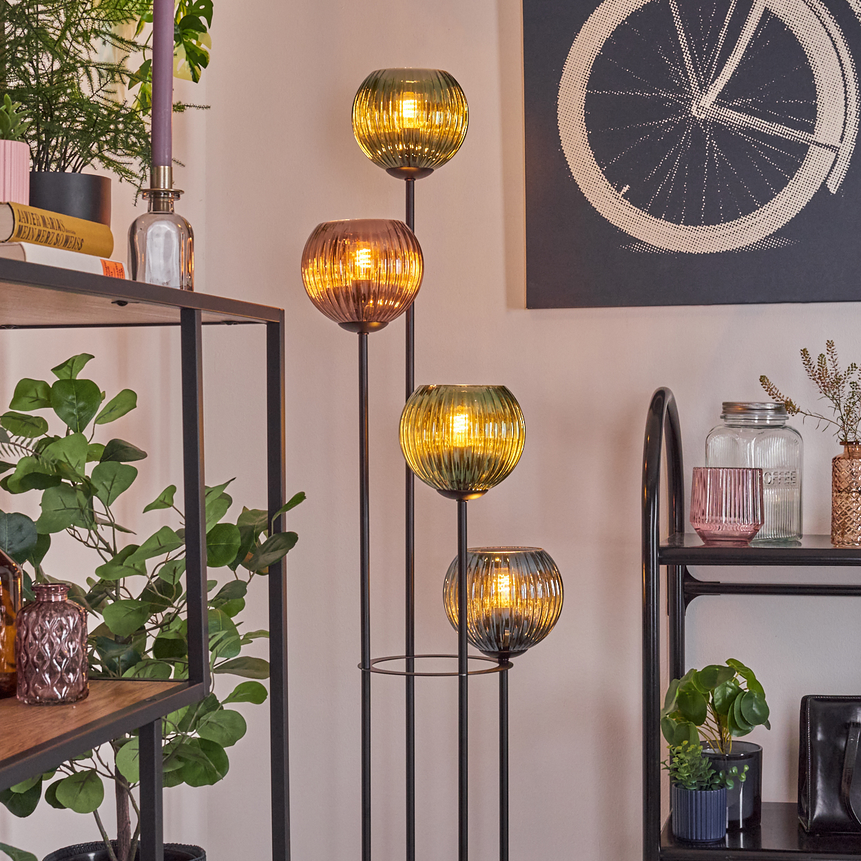 Dekorative Stehlampe mit vier Glaskugeln. Regale halten Pflanzen, Bücher und Gläser. Ein Fahrradrad-Kunstwerk hängt an der Wand.