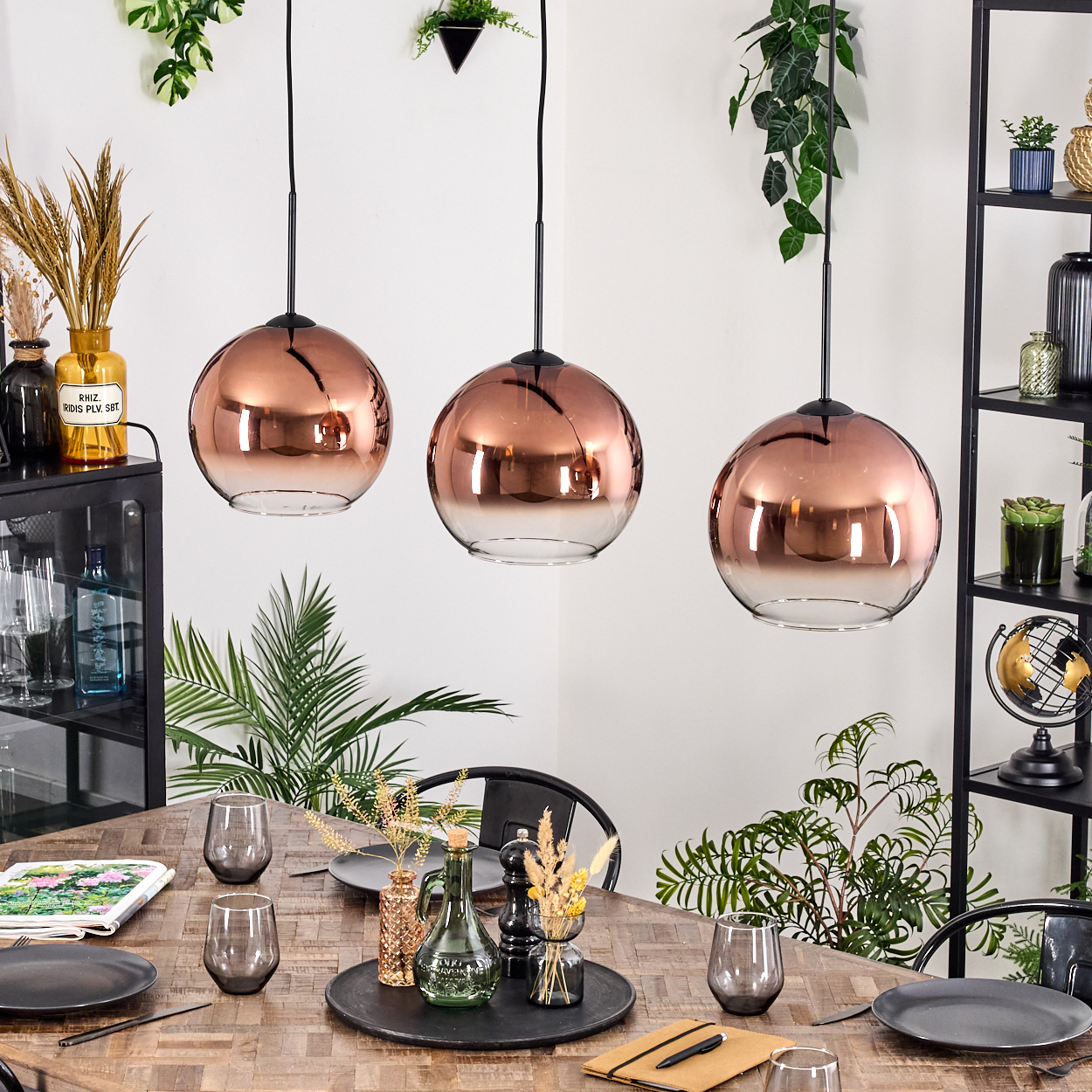 Esszimmer mit drei kupferfarbenen Pendelleuchten. Tisch mit Tellern, Gläsern und Dekorationsartikeln. Pflanzen und Regale im Hintergrund.