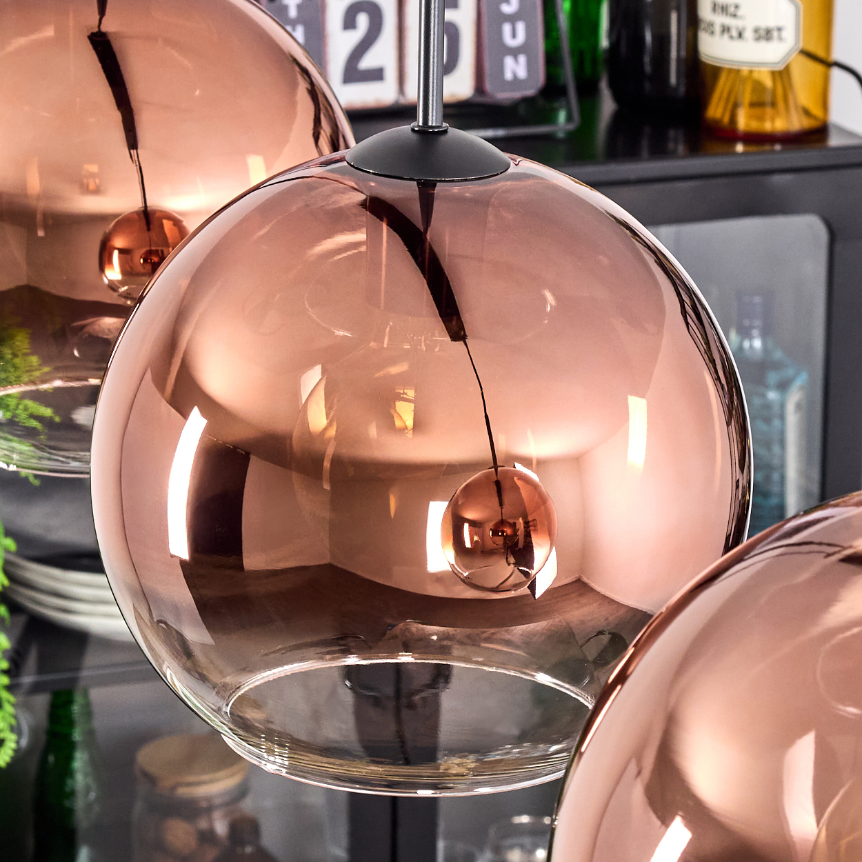 Drei kupferfarbene Glas-Pendelleuchten hängen. Im Hintergrund sind ein Kalender und Flaschen zu sehen, die sich im Glas spiegeln.