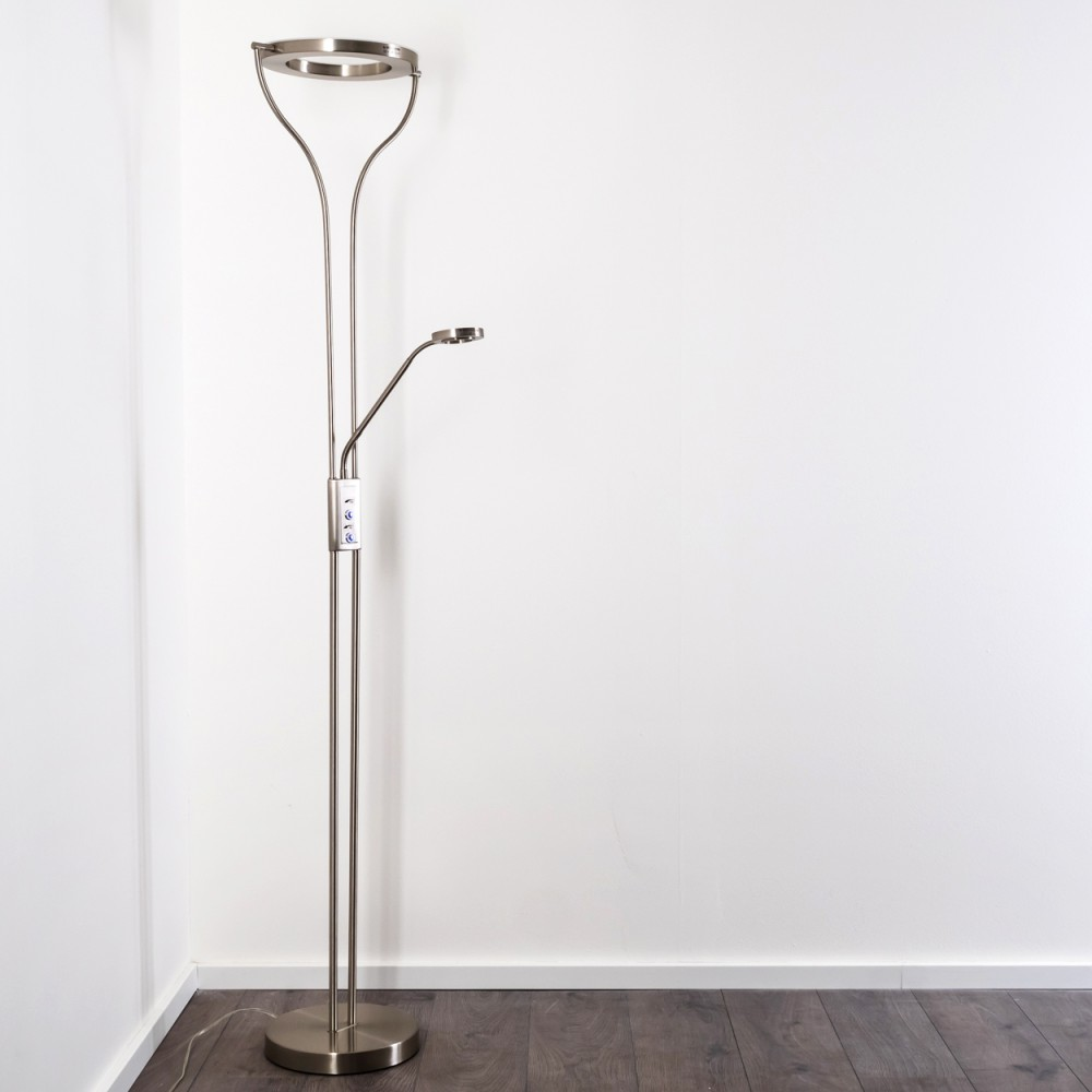 Eine moderne, silberne Stehlampe steht vor einer weißen Wand und einem dunklen Holzboden. Sie hat eine kreisförmige Oberseite und ein kleineres Licht.