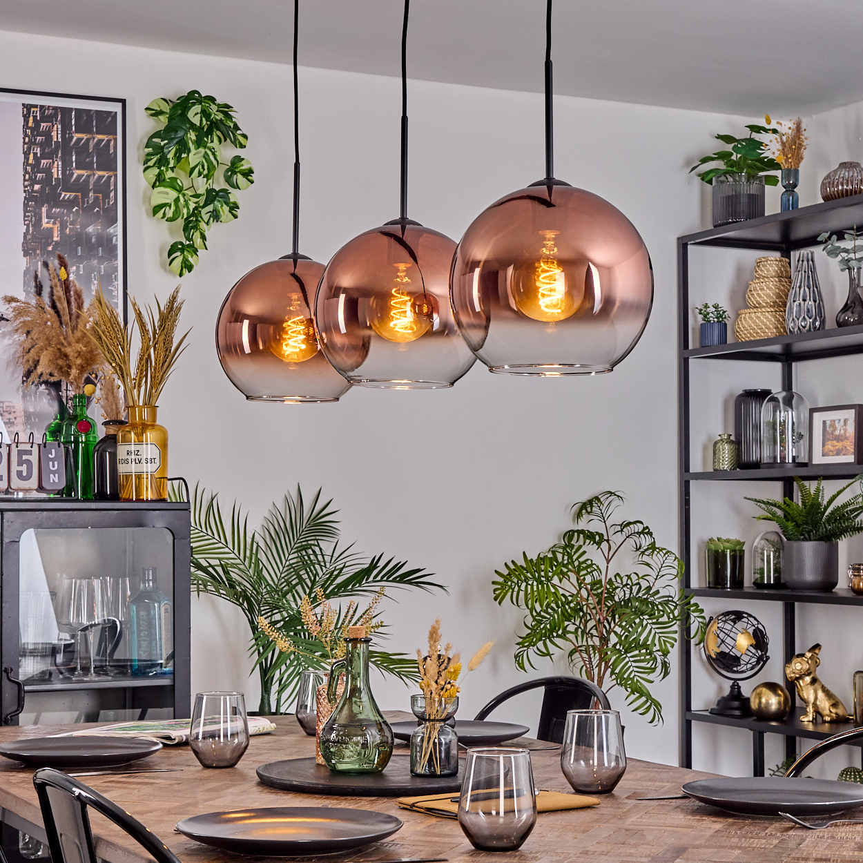 Esszimmer mit Holztisch, drei Hängelampen und verschiedenen Dekorationen, darunter Pflanzen und Regale.