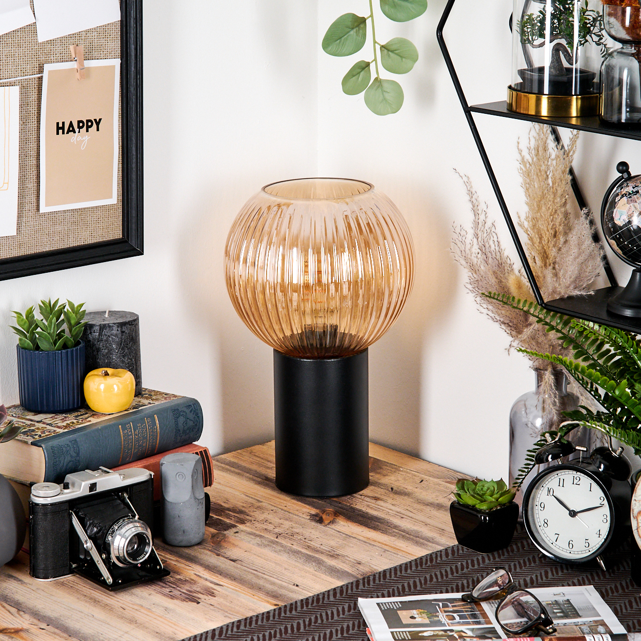 Eine Schreibtischecke mit Lampe, Büchern, Kamera und Uhr. Eine Notiz mit Happy Day ist an der Wand befestigt. Pflanzen und Dekoration sind ebenfalls vorhanden.