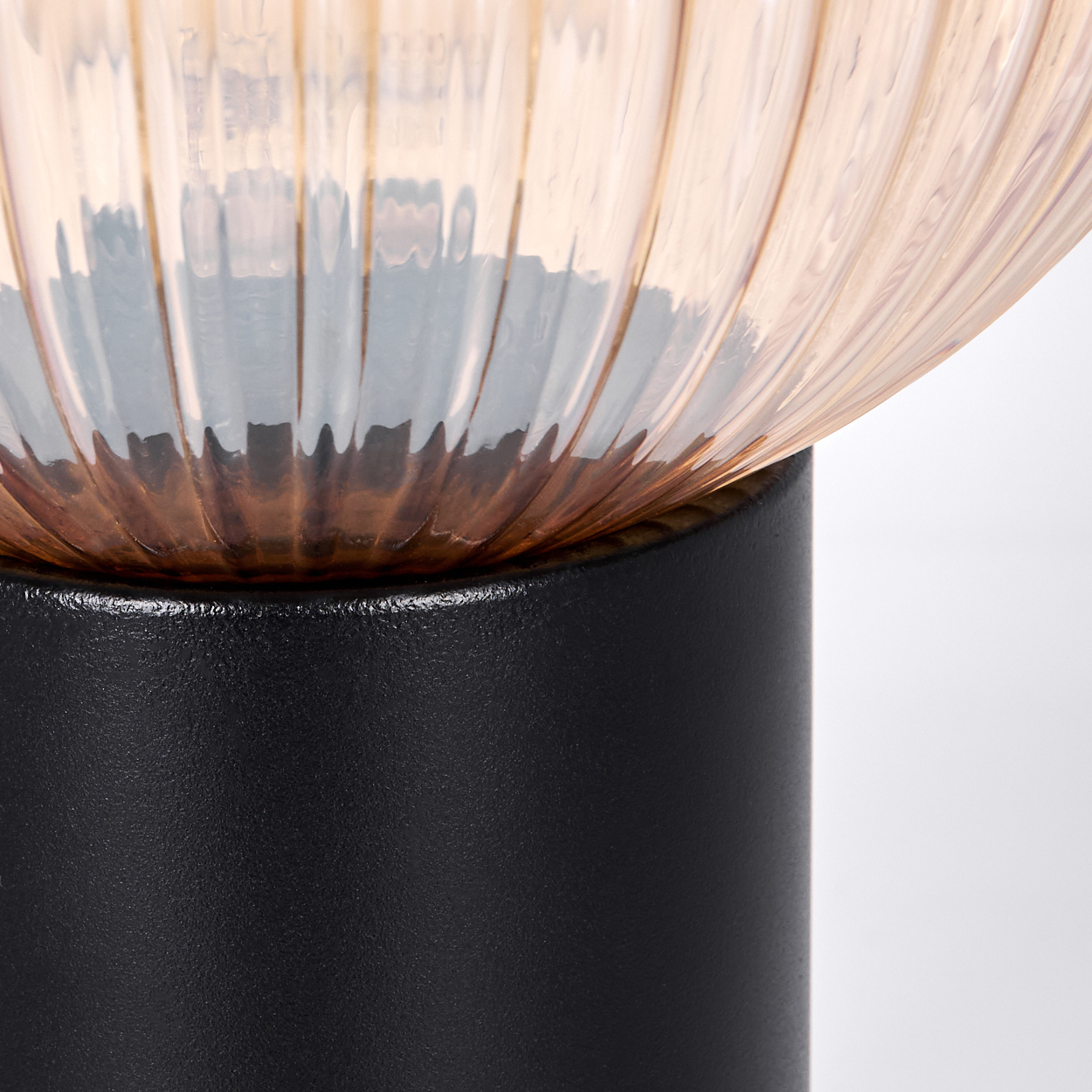 Nahaufnahme einer Lampe. Die Oberseite ist ein geripptes, pfirsichfarbenes Glas. Sie sitzt auf einem schwarzen zylindrischen Sockel vor einem weißen Hintergrund.