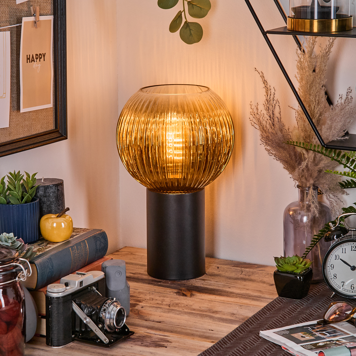 Eine dekorative Lampe mit geripptem, bernsteinfarbenem Glasschirm, schwarzem Sockel und brennender Glühbirne steht auf einem Holztisch.