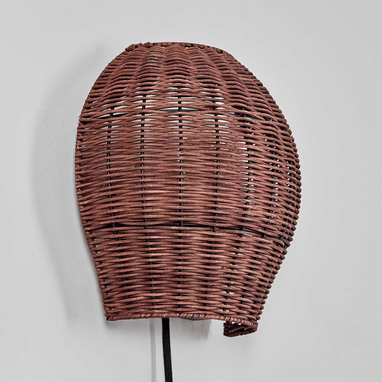 Eine gewebte braune Lampe hängt an einer weißen Wand. Sie ist oval und hat ein schwarzes Kabel. Die Webart ist detailliert sichtbar.