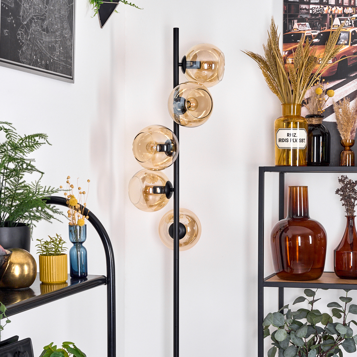 Eine moderne Stehlampe mit fünf bernsteinfarbenen Glaskugeln. Regale zeigen Vasen und Pflanzen. Eine Karte und ein Stadtfoto an der Wand.