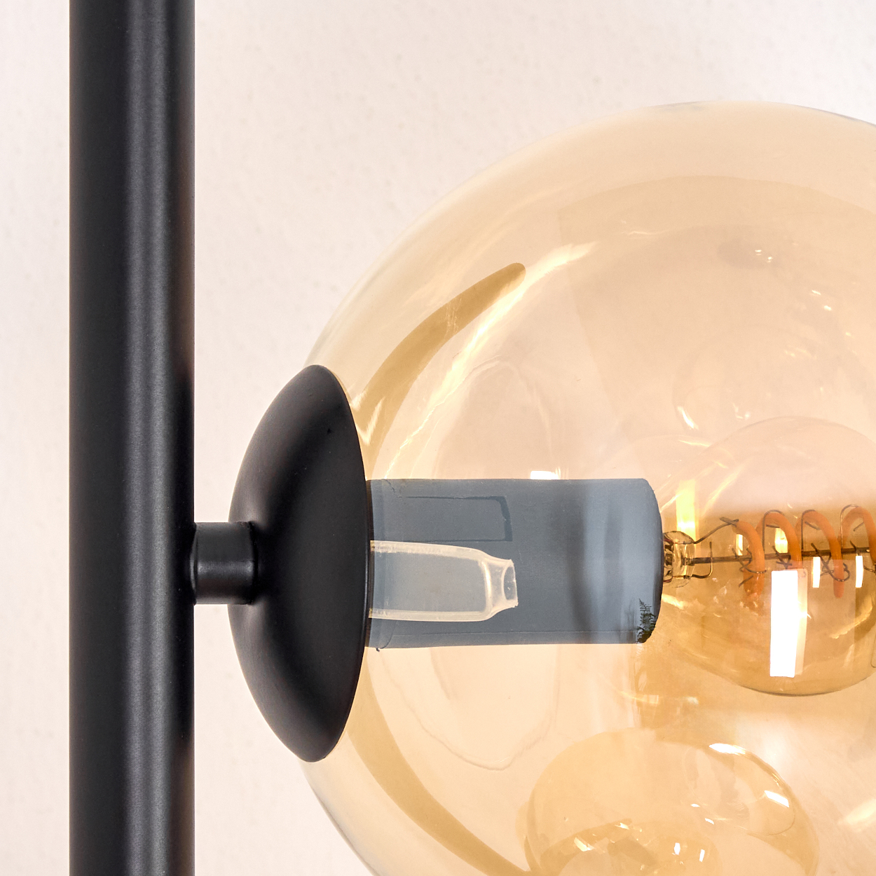Nahaufnahme einer Lampe. Ein schwarzer Metallrahmen trägt eine große, bernsteinfarbene Glaskugel. Der glühende Glühfaden der Glühbirne ist sichtbar.