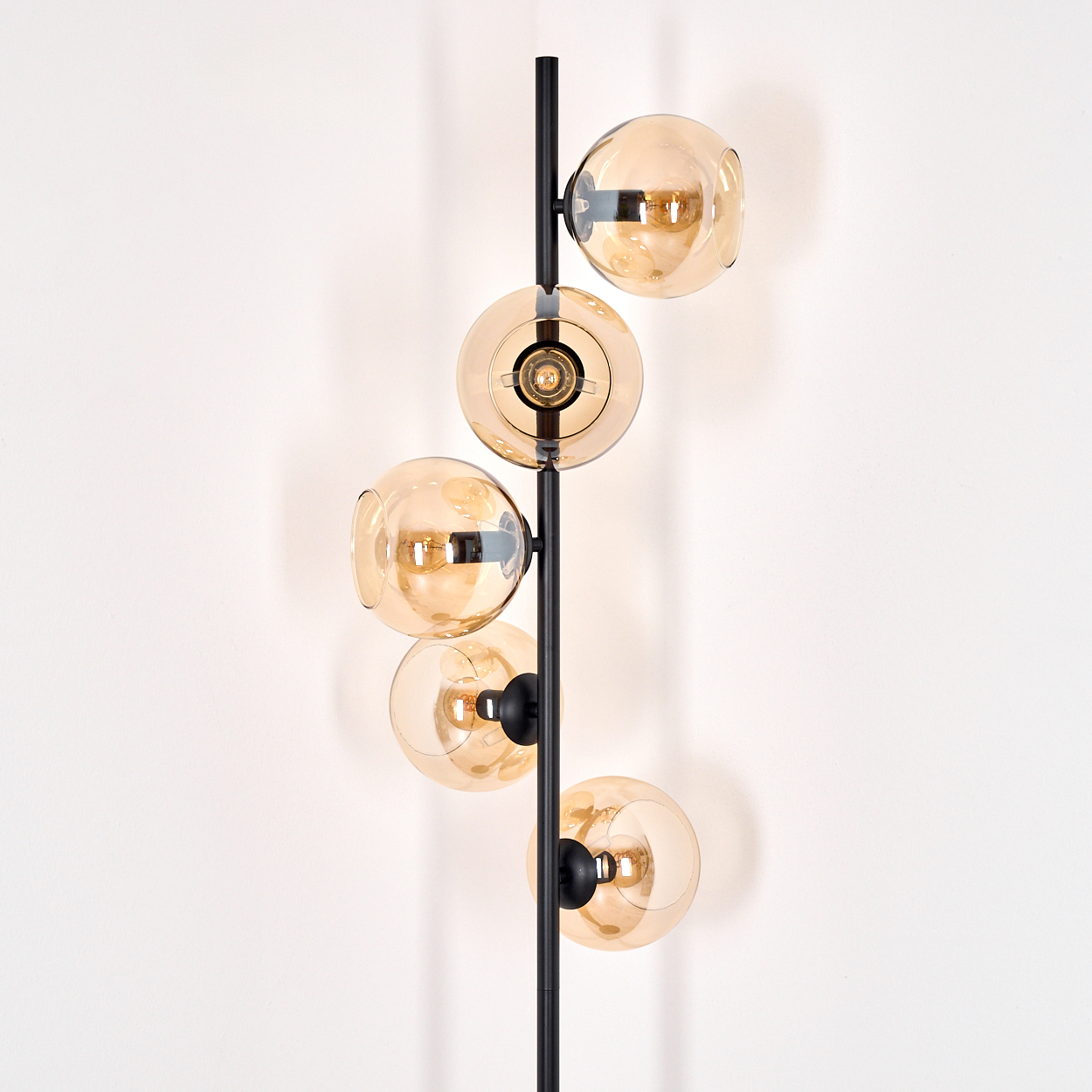 Eine moderne Stehlampe mit fünf kugelförmigen, bernsteinfarbenen Glasschirmen. Die Lampe hat ein schwarzes Metallgestell und steht vor einer weißen Wand.