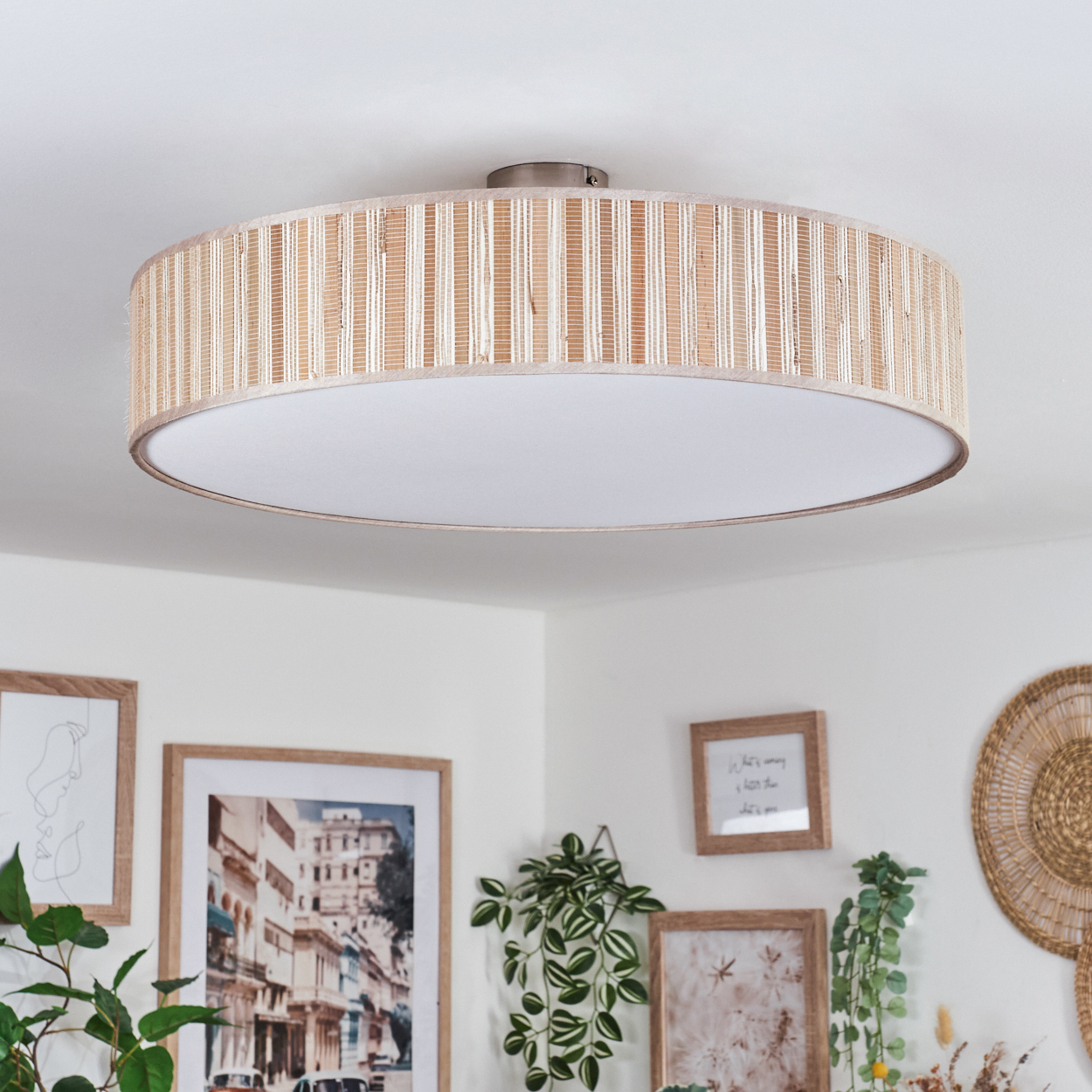 Eine runde Deckenlampe mit gewebter Struktur. Darunter dekorieren gerahmte Bilder und Pflanzen die weiße Wand. Auch eine Wanddekoration aus Weide ist vorhanden.