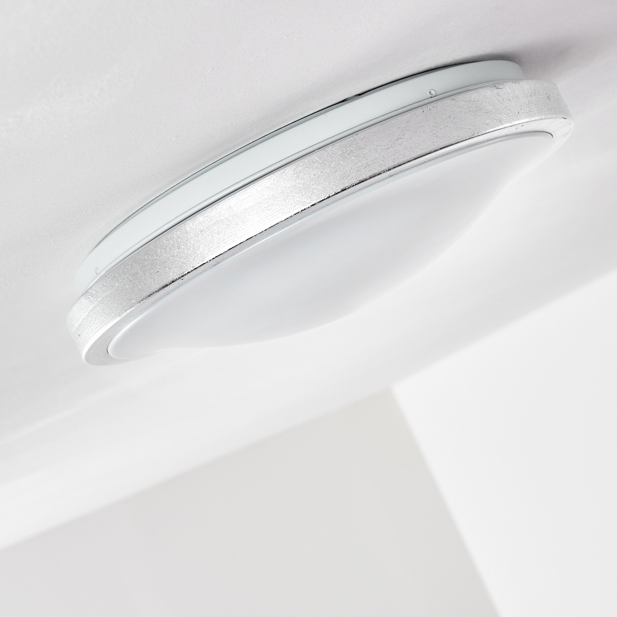 Nahaufnahme einer weißen Deckenleuchte mit silbernem Rand und weißem, abgerundeten Diffusor. Der Hintergrund ist eine weiße Decke.