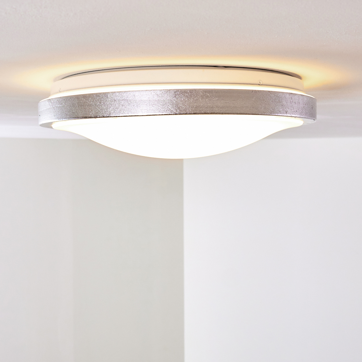 Eine moderne Deckenleuchte mit silbernem Band um einen weißen, gebogenen Diffusor, beleuchtet. Es ist vor weißen Wänden und Decke.