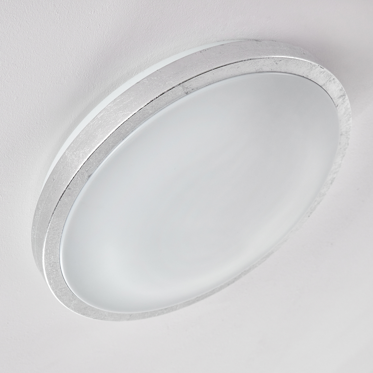 Nahaufnahme einer Deckenleuchte. Sie hat einen silbernen Rahmen und eine weiße, matte Glasabdeckung. Der Hintergrund ist eine weiße Decke.