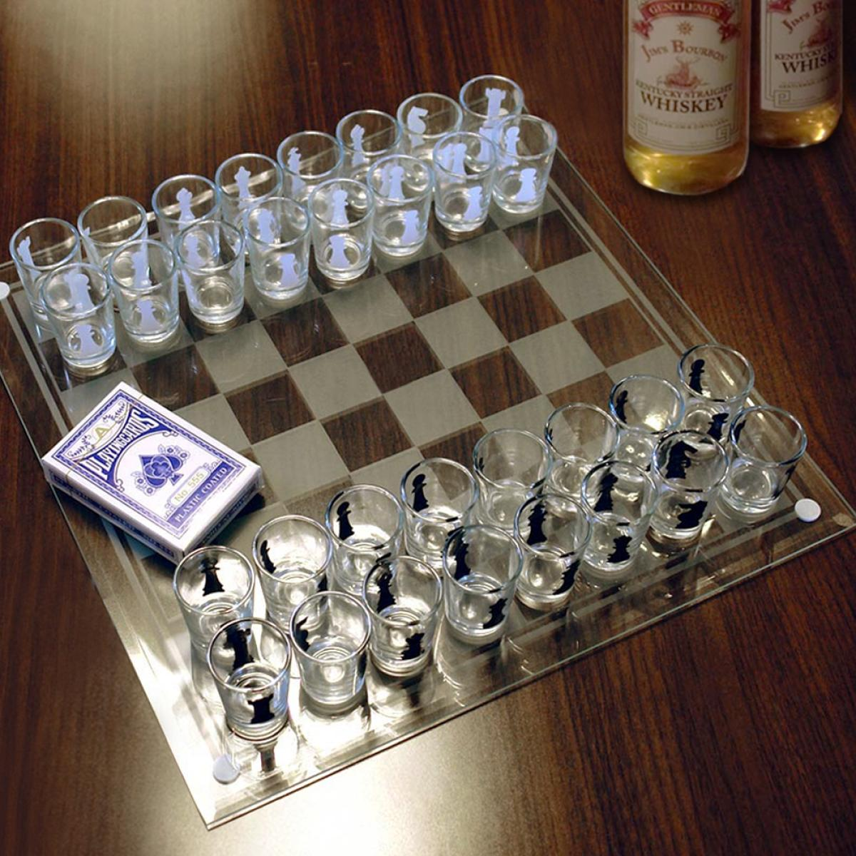 Een schaakbord van glas met borrelglaasjes als schaakstukken. Een pak kaarten en whiskyflessen staan op de houten tafel.