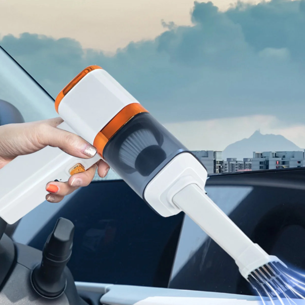 Eine Hand hält einen weiß-orangenen Handstaubsauger und reinigt das Innere eines Autos. Im Hintergrund sind Berge und Wolken zu sehen.