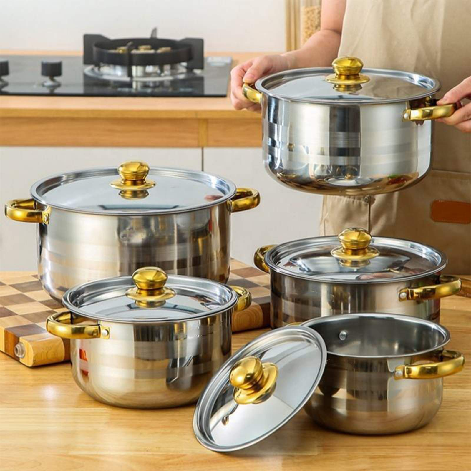 Edelstahltöpfe mit goldenen Griffen und Deckeln stehen auf einem Holztisch. Eine Person hält einen Topf.