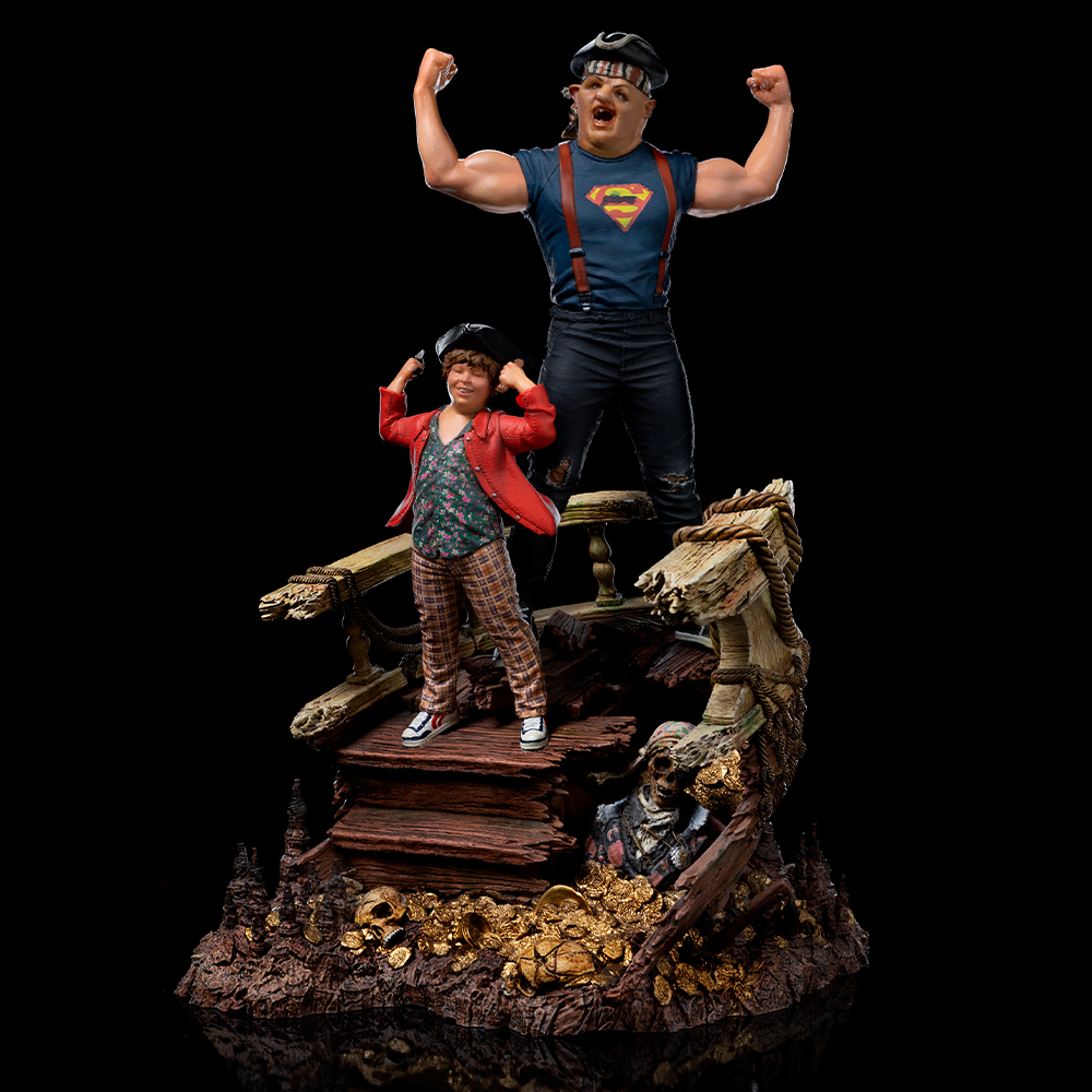 Statue von zwei Figuren, die sich biegen. Einer trägt ein Superman-Shirt, der andere eine rote Jacke. Sie stehen auf einer holzgefüllten Struktur.
