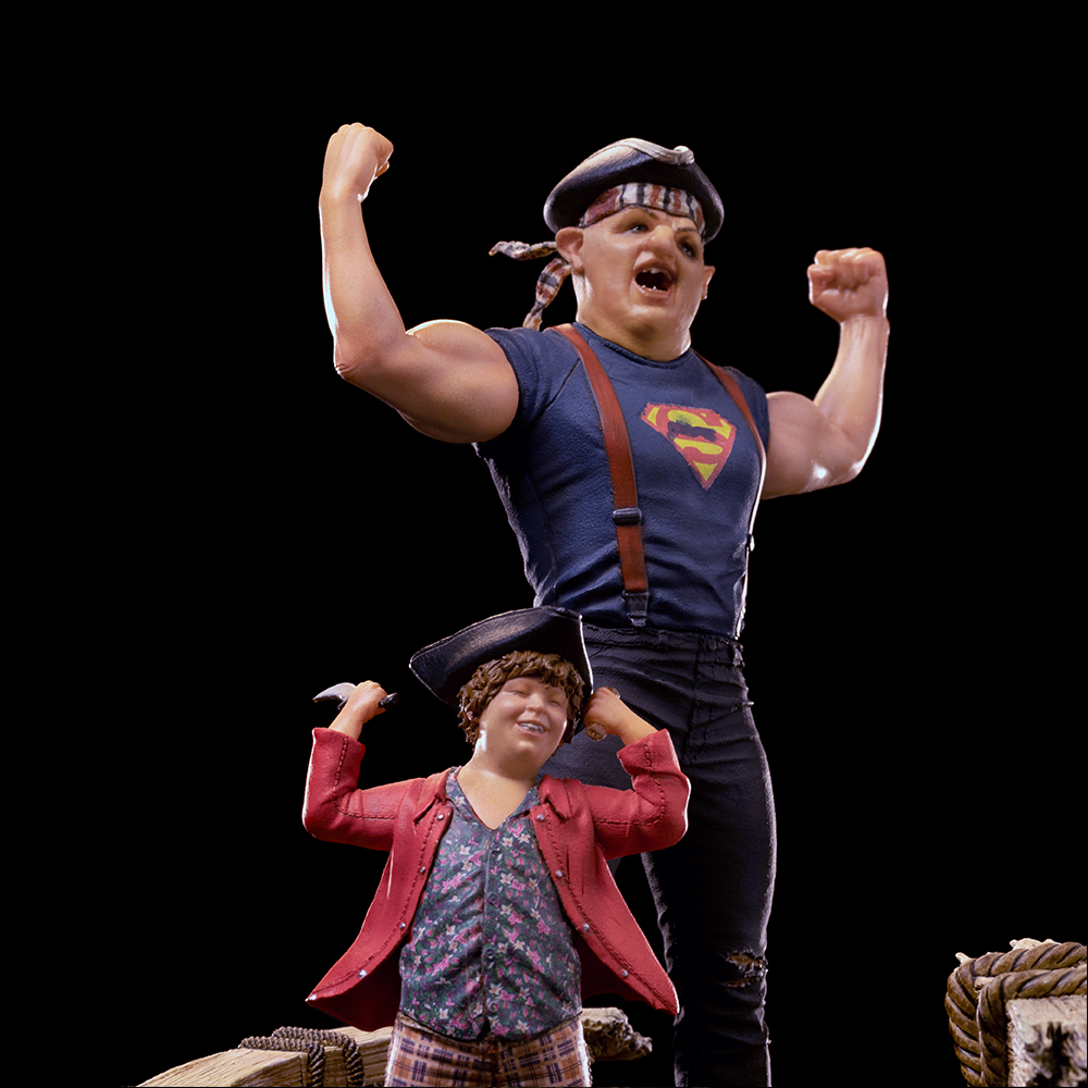 Statuen von zwei Figuren, eine muskulös mit Superman-Shirt, die andere kleiner mit roter Jacke, beide heben Arme, schwarzer Hintergrund.